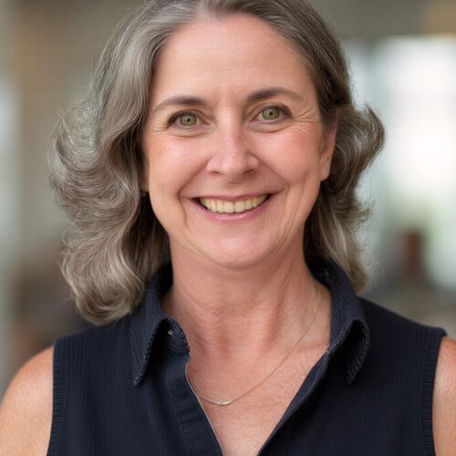 woman with gray wavy hair in a black sleeveless dress smiling inside an office building