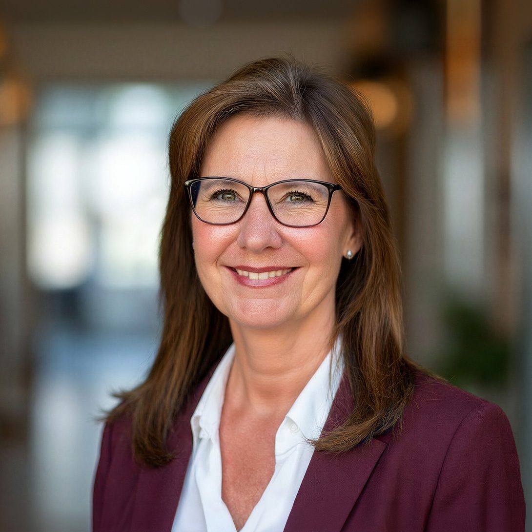 businesswoman with brunette hair and glasses in a burgundy suit smiling indoors