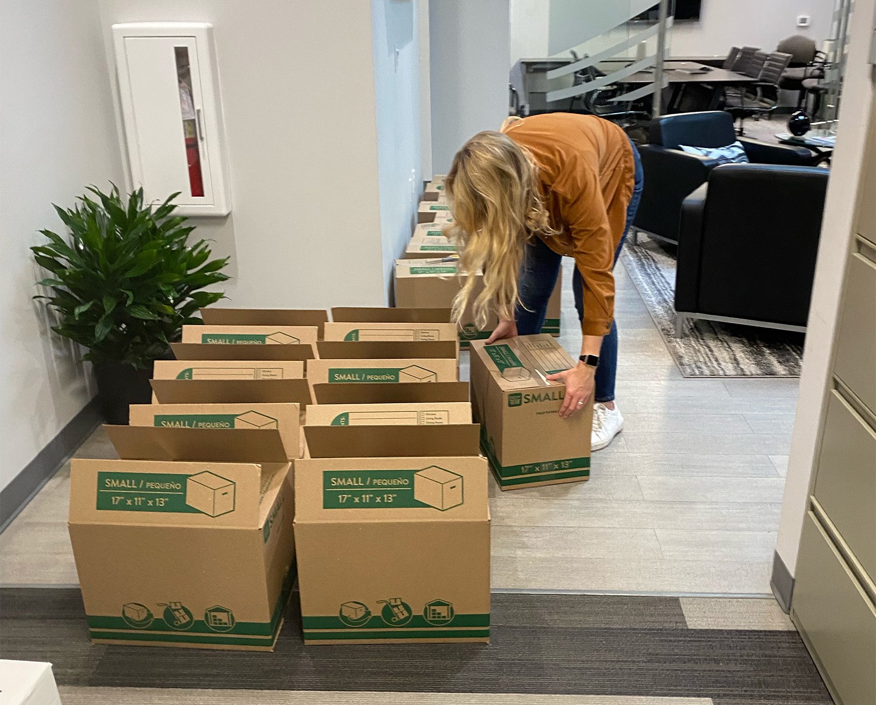 blond woman bending over to tape up one box of many on the floor