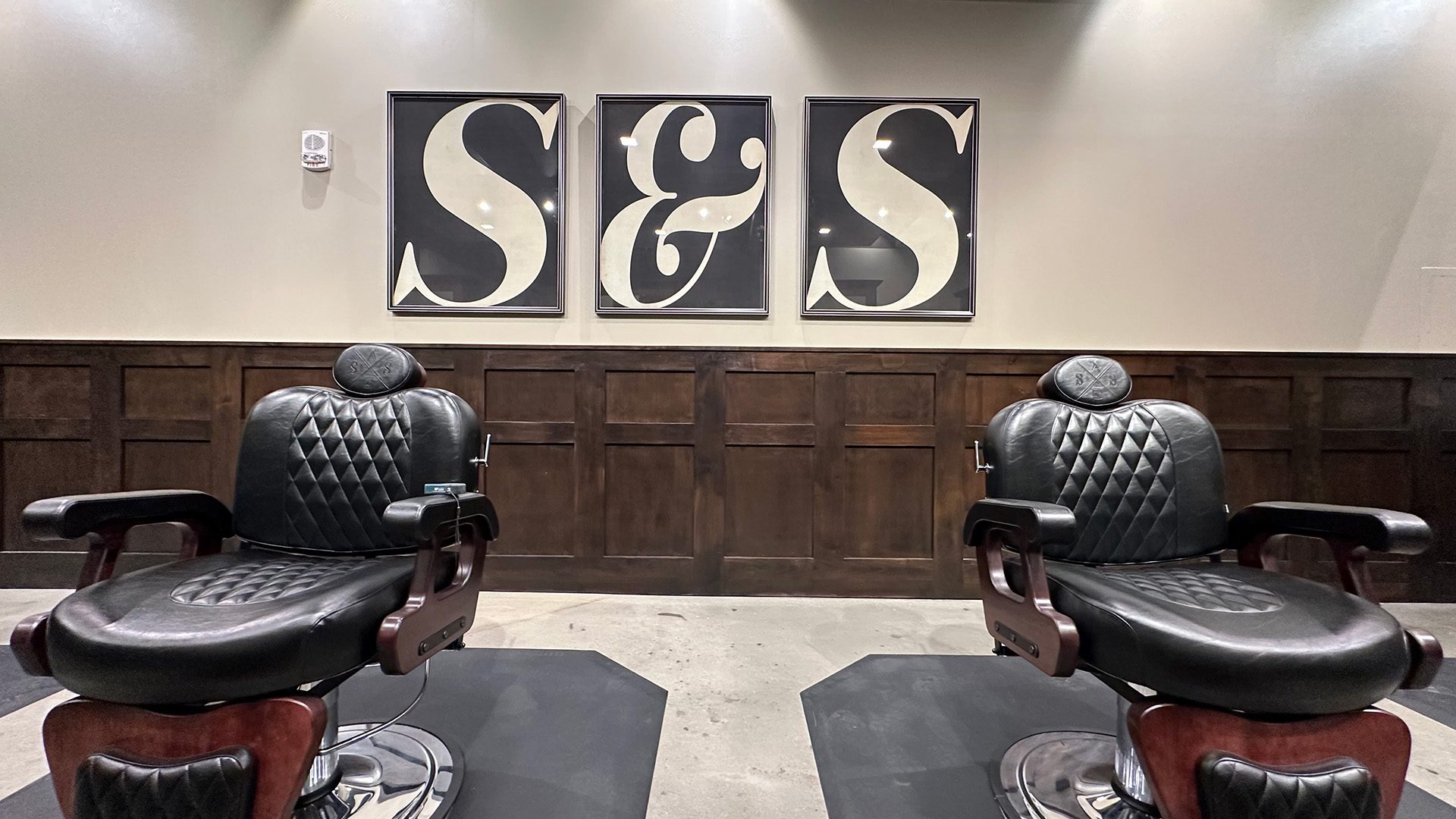 'S&S' art on the wall with wood wainscoting and two leather barber chairs