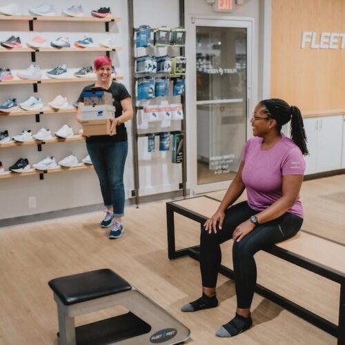 interior of Fleet Feet store with two women, one sales associate helping a seated woman find running shoes