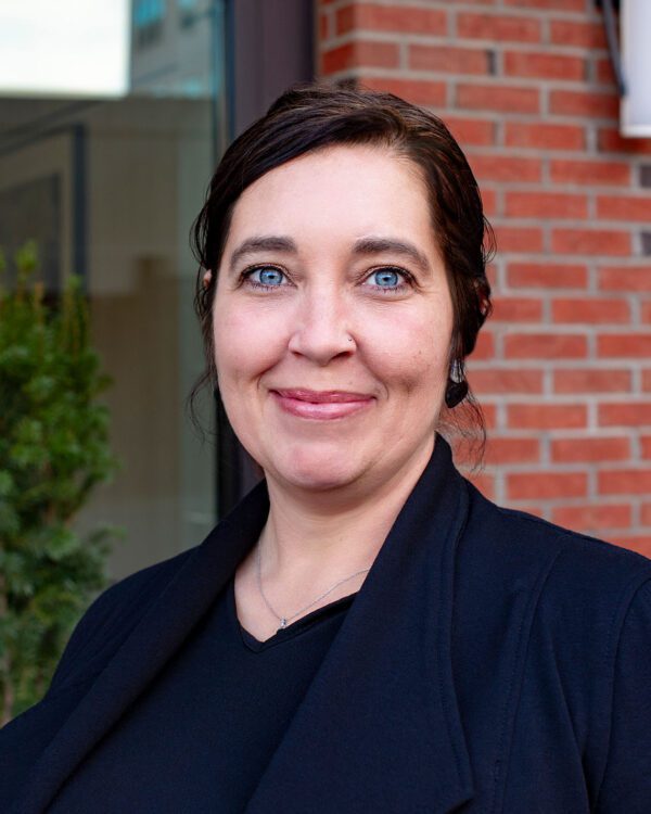 Amy Davis woman smiling with dark hair and a black blazer in front of window outside