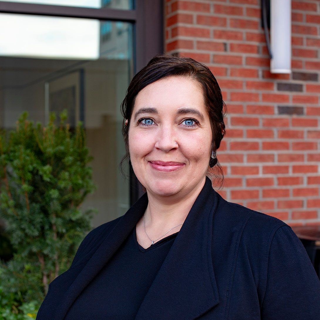 Amy Davis woman smiling with dark hair and a black blazer in front of window outside